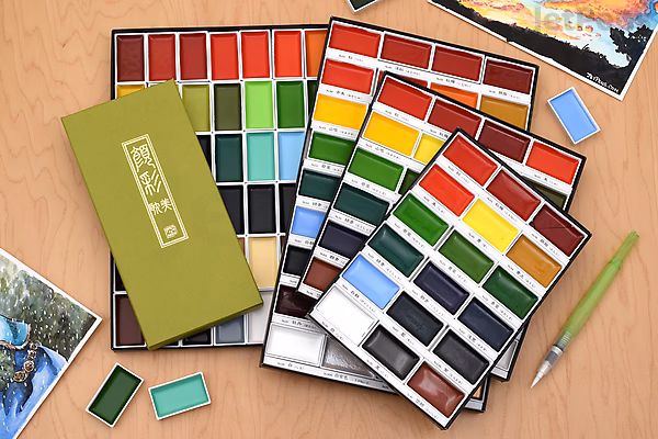 Kuretake Gansai Tambi 12-color watercolor palette set with four open trays showing vibrant colors, a closed olive green box, two paintbrushes, and two small watercolor pans on a wooden surface.