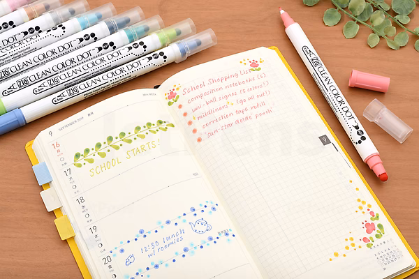 Open notebook with colorful dot patterns and handwritten notes, surrounded by Kuretake Zig Clean Color Dot Double-Sided Markers in metallic colors, including pink, blue, and green, on a wooden surface with a small plant branch nearby.