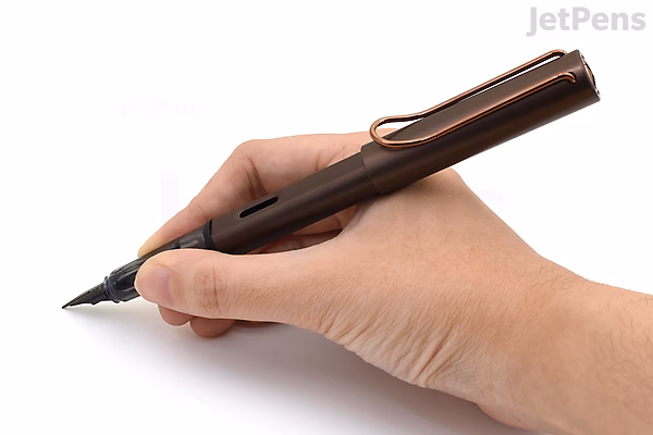 A hand holding a LAMY Lx Marron fountain pen with an extra fine nib, poised for writing. The pen has a sleek, dark brown metallic finish with a clip on the cap.