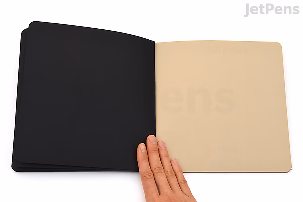 An open Stillman & Birn Nova Sketchbook with softcover, measuring 7.5 by 7.5 inches. The left page is black, and the right page is a light beige color. A hand is holding the bottom edge of the right page.