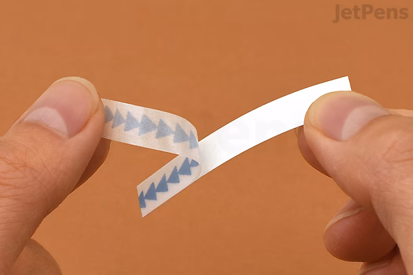 A close-up of two hands peeling a strip of King Jim Kitta Washi Tape with a blue airmail pattern of small triangles on a white background.