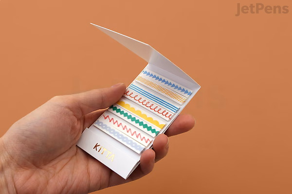 A hand holding a small booklet of King Jim Kitta Washi Tape featuring colorful airmail-themed designs, including stripes, arrows, and envelopes.