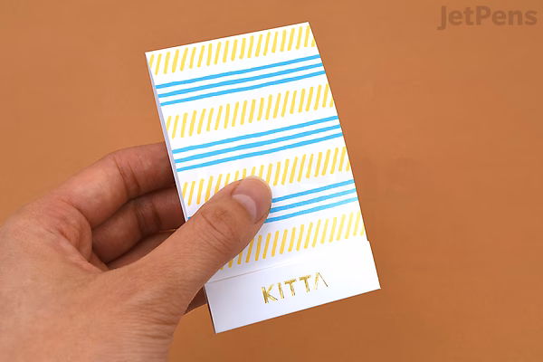 A hand holding a small rectangular sheet of King Jim Kitta Washi Tape with an airmail design featuring diagonal blue and yellow stripes on a white background.