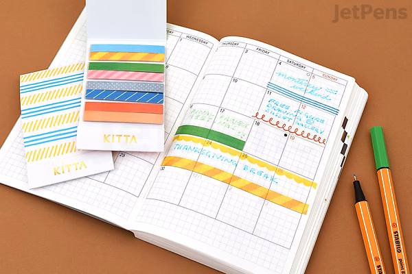 King Jim Kitta Washi Tape - Airmail design shown with colorful patterned tape strips applied in a planner, alongside two orange pens on a brown surface.