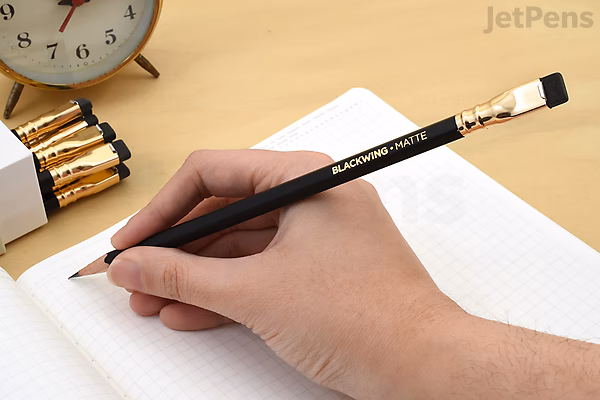Hand holding a Blackwing 602 pencil writing in a notebook with a clock and a pack of 12 Blackwing 602 pencils in the background.