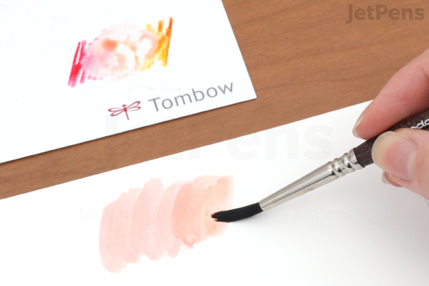 A hand holding a paint brush, applying light-colored ink to paper. The ink came from two brighter colors of PITT Pen ink that were applied to a palette from the Tombow Dual Brush Pen Blending Kit.