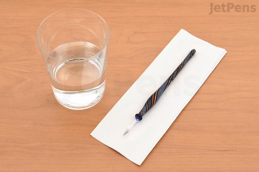 A glass of water shown next to a glass dip pen drying on a paper towel.