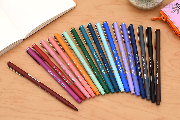 A set of 18 Marvy Le Pen Flex brush pens arranged in a row on a wooden surface, displaying a gradient of colors from red, pink, orange, yellow, green, blue, purple to black. The pens have colored caps matching the ink color.