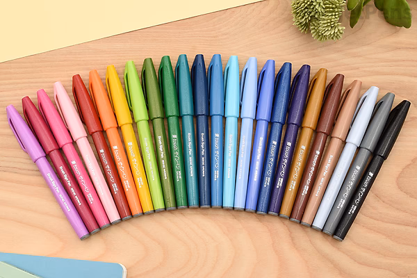 Set of 12 Pentel Fude Touch Brush Sign Pens arranged in a row on a wooden surface, showcasing a range of vibrant colors from pinks and reds to blues, browns, and black. Some green leaves are partially visible at the top.