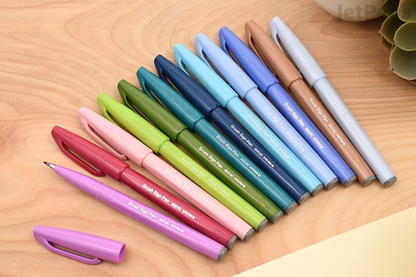 A set of twelve Pentel Fude Touch Brush Sign Pens arranged in a fan shape on a wooden surface. The pens display a gradient of colors including shades of pink, red, green, blue, purple, and gray, with one pen cap removed and placed nearby.