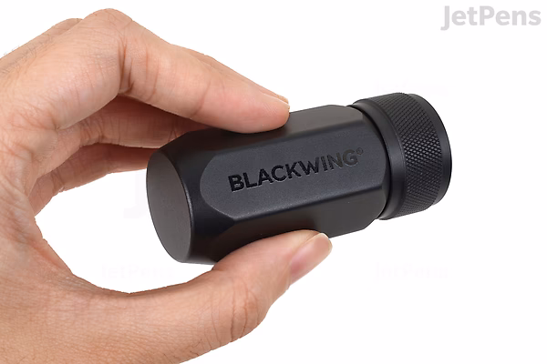 A hand holding a small black cylindrical container labeled "BLACKWING," designed to store pencils or leads.