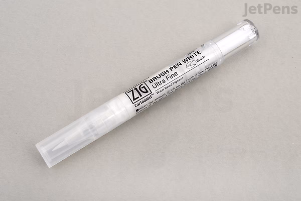 A close-up photo of a Kuretake ZIG Cartoonist Brush Pen with white ink and an ultra fine tip, lying on a light gray surface. The pen has a transparent cap and a white body with black text.