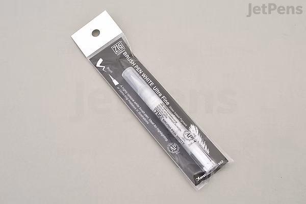 A packaged Kuretake ZIG Cartoonist Brush Pen with white ink and an ultra-fine tip, displayed on a gray surface. The pen is in a clear plastic blister pack with a black and white cardboard backing.