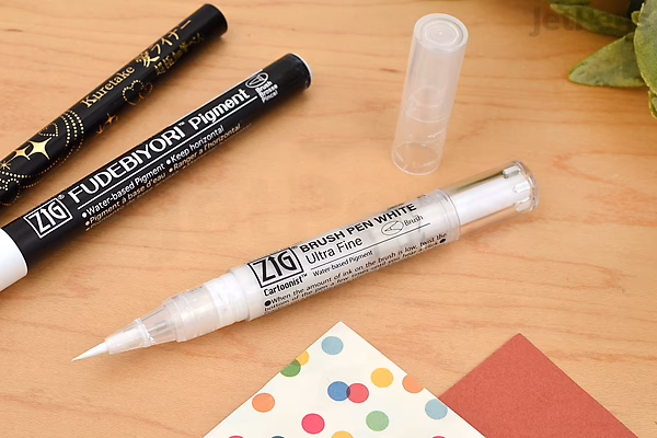 A Kuretake ZIG Cartoonist Brush Pen with white ink and an ultra-fine tip lies uncapped on a wooden surface, alongside two other black pens and colorful paper pieces.