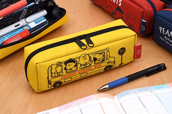 Yellow rectangular pencil case with black zipper featuring Peanuts characters riding a school bus, shown open with pens inside, placed on a desk with notebooks and other pencil cases nearby.