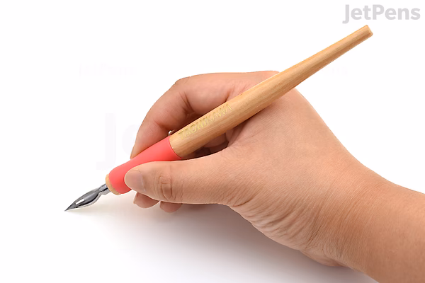 A hand holding a Tachikawa T-36 nib holder with a wooden body and a pink rubber grip, fitted with a metal calligraphy nib.
