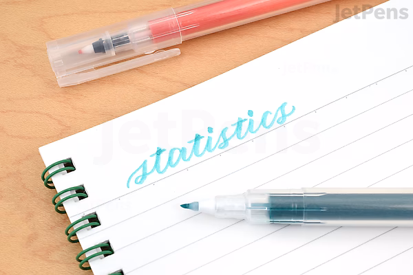 Close-up of a Kuretake Karappo empty felt tip pen with a fine wick tip, uncapped and lying on a notebook with green spiral binding. The pen is writing the word "statistics" in light blue ink on lined paper.