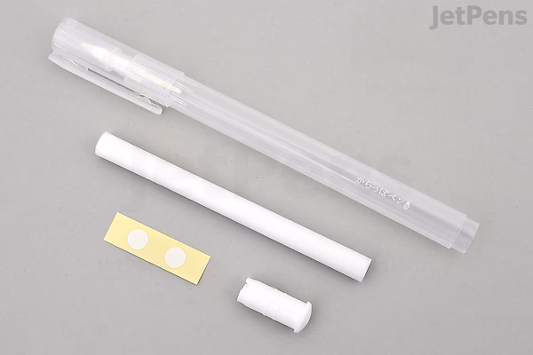 A disassembled Kuretake Karappo Empty Brush Pen with Wick in fine size, showing the clear plastic pen body, white ink reservoir, small white cap, and two round adhesive pads, all laid out on a gray surface.