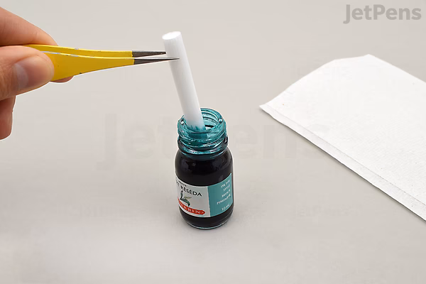 A hand holding a yellow-handled Kuretake Karappo empty brush pen with a wick tip, dipping it into a small bottle of teal ink beside a folded white paper towel.
