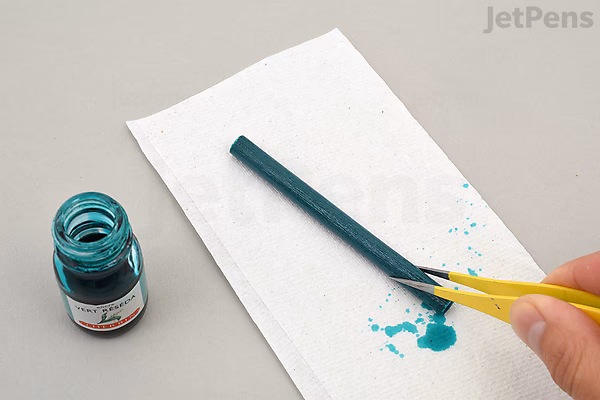 A person using scissors to cut the wick of a Kuretake Karappo empty brush pen, placed on a white paper towel next to an open bottle of teal ink.