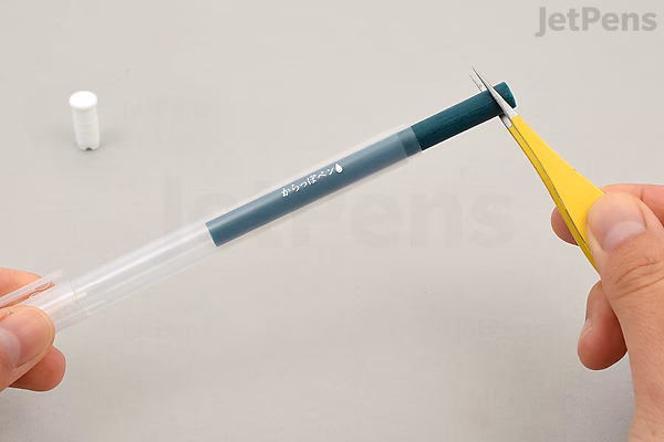 A close-up photo of a person holding a yellow utility knife and cutting the wick inside a transparent Kuretake Karappo empty brush pen with a fine tip. The pen's cap is placed on the surface nearby.