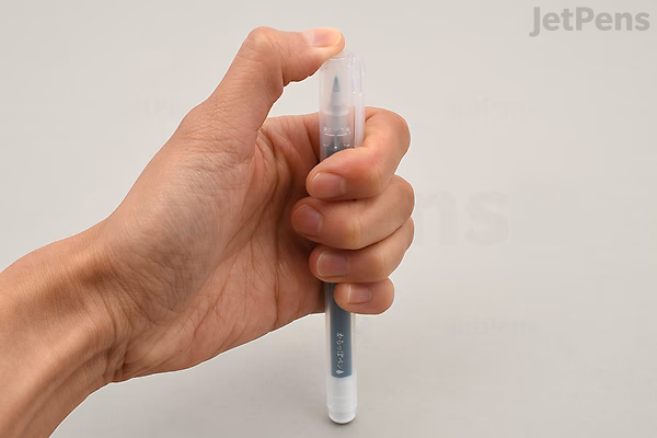 A hand holding a Kuretake Karappo empty brush pen with a fine wick tip against a plain light background.