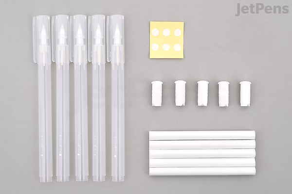 Five clear Kuretake Karappo empty brush pens with white caps, five white replacement wicks, five white inner pen barrels, and a small yellow square with six white dots, all arranged neatly on a light gray background.