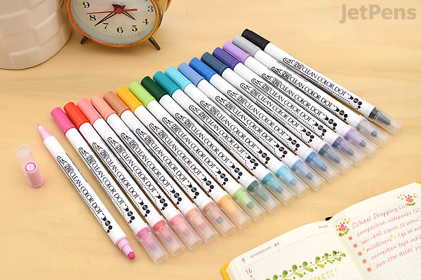 Set of 12 Kuretake ZIG Clean Color Dot double-sided markers arranged on a desk with a clock, notebook, and cup in the background. Each marker has a round dot tip on one end and a brush tip on the other.
