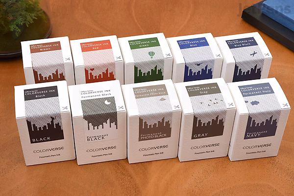 Twelve small white boxes of Colorverse Black Ink, each labeled with different colors and arranged in three rows on a wooden surface. Each box contains a 30 ml bottle of ink.