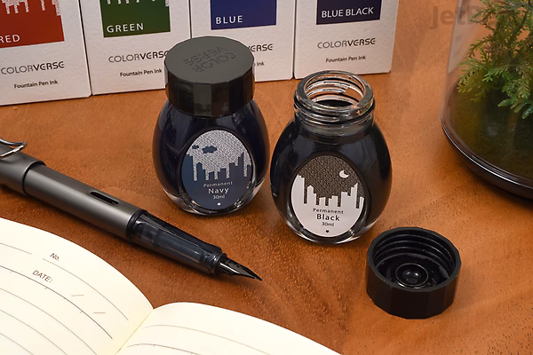 Two small bottles of Colorverse Black Ink labeled "Office," one open with the cap beside it, and a black fountain pen resting on an open notebook on a wooden surface.