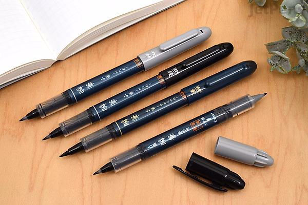 Three Pilot Shunpitsu Pocket Brush Pens in black and gray, shown with caps removed and placed nearby, resting on a wooden surface next to an open notebook and a small plant. The pens have double-sided tips.