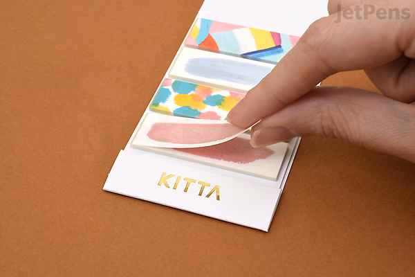 A hand is peeling a strip of King Jim Kitta Washi Tape from a small, white card labeled "KITTA." The tape features colorful, abstract patterns in pastel shades. The card holds multiple strips of decorative tape arranged vertically.