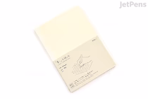 A Midori MD Notebook in A5 size with lined pages, shown closed with a cream-colored cover and a beige paper band around it featuring minimalist text and a simple line drawing of a hand holding a pen.