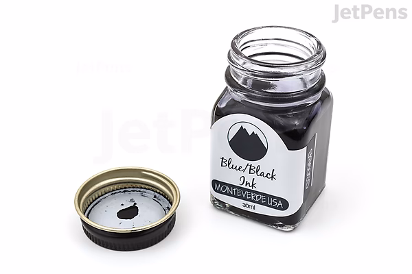 A 30 ml glass bottle of Monteverde Black Ash Ink with its gold-colored screw cap removed and placed beside it, showing the dark ink inside. The bottle has a white label with black text and a black circle design.