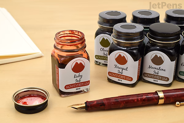 Open bottle of Monteverde Black Ash Ink with cap off, surrounded by other Monteverde ink bottles and a red fountain pen on a wooden desk.