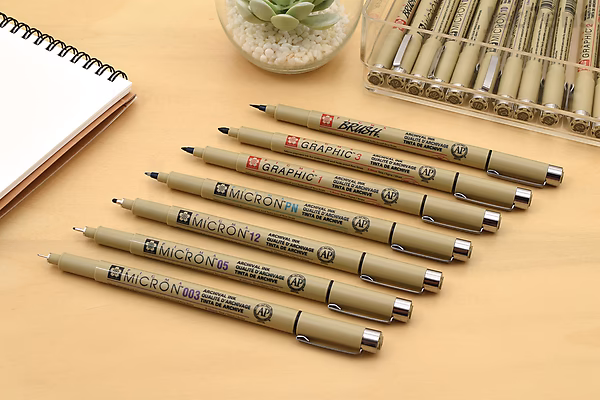 Eight Sakura Pigma Micron pens arranged on a wooden surface next to a notebook and a small plant. The pens are size 02 (0.3 mm) with black ink.