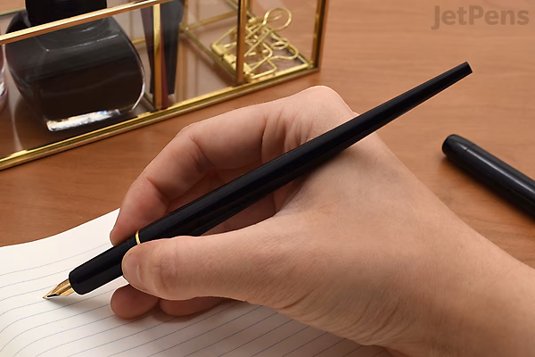 A hand holding a red Platinum Desk Fountain Pen with an extra fine nib, writing on lined paper. The pen has a sleek, modern design with a black grip section. In the background, there is a glass ink bottle and a gold-colored pen holder.