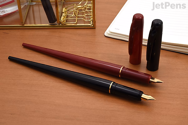Two Platinum Desk Fountain Pens with extra fine nibs, one in red and one in black, resting on a wooden desk near a notebook and a decorative gold item. Caps are removed and placed upright behind the pens.