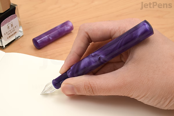 A hand holding a purple marbled Majohn N6 fountain pen with an extra fine fude nib, positioned above a white surface. The pen cap lies nearby, and a bottle of purple ink is visible in the background.