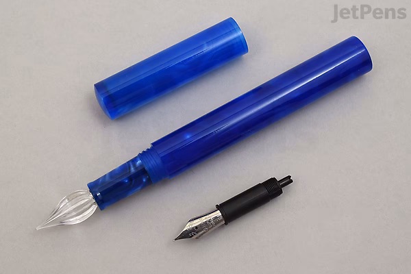 A Majohn N6 fountain pen in iris blue swirl color with its cap removed, showing a clear glass dip nib attached to the pen body. Next to it lies an extra fine fude nib with a black grip section. The pen and nibs are placed on a light gray surface.