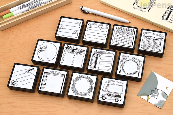 A collection of 11 small square pre-inked stamps with various designs, including calendars, clocks, and decorative frames, arranged on a wooden surface next to pens and a small card with a bird illustration.
