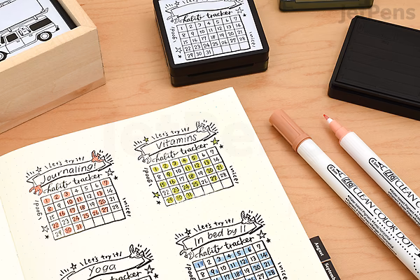 A planner page stamped with colorful bird-themed calendar stamps in red, yellow, blue, and green. Nearby are a wooden stamp box, a black ink pad, and two dual-tip markers in beige and white.
