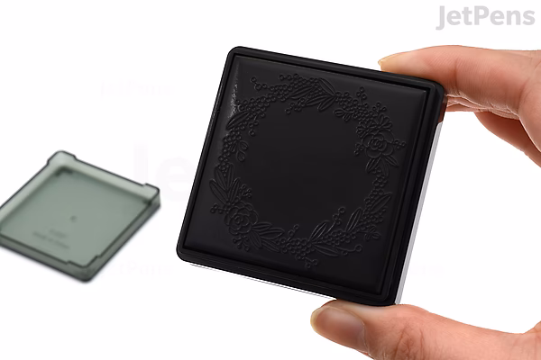 A hand holding a black Midori paintable pre-inked stamp with a wreath design, next to its translucent protective cover.