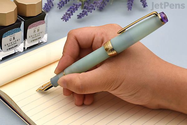 A hand holding a Sailor Pro Gear Slim Shikiori fountain pen in a soft green color with gold accents, poised to write on a lined notebook. The pen has a 21k gold medium-fine nib. In the background, there are two ink bottles and some lavender flowers.