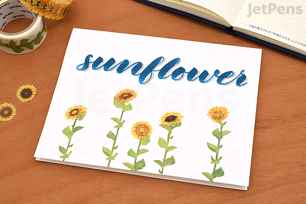 A sheet with five hand-drawn sunflower illustrations in various stages of bloom, labeled "sunflower" in blue cursive at the top, placed on a wooden desk with a small jar of tape and a notebook nearby.