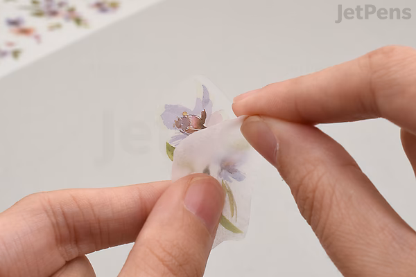 A person holding a small floral print-on transfer with both hands, preparing to apply it onto a surface. The transfer features delicate purple and white flowers.