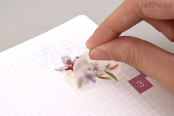 A hand holding a small, translucent flower-shaped wooden applicator with a printed floral design, positioned above a grid-lined sheet of paper.