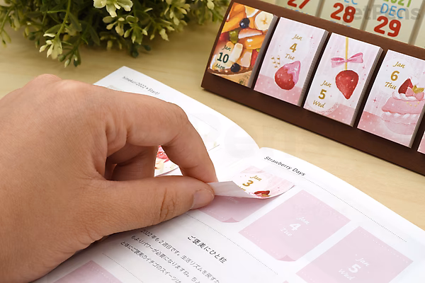 A hand peeling a small pink sticky note from a Himekuri Sticky Note Calendar 2022 booklet, with a wooden calendar stand displaying daily sticky notes in the background. A plant is partially visible on the side.