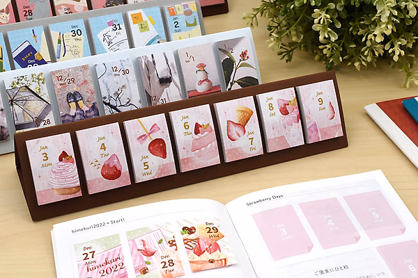 A Himekuri Sticky Note Calendar 2022 displayed on a wooden stand, featuring daily illustrated sticky notes with cute designs. An open booklet with matching pink-themed pages lies in front, surrounded by other stationery items and a green plant.