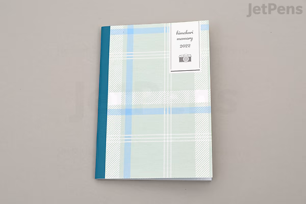 A closed Himekuri Sticky Note Calendar 2022 & Booklet with a blue and green plaid cover and a teal spine, lying on a light gray surface.